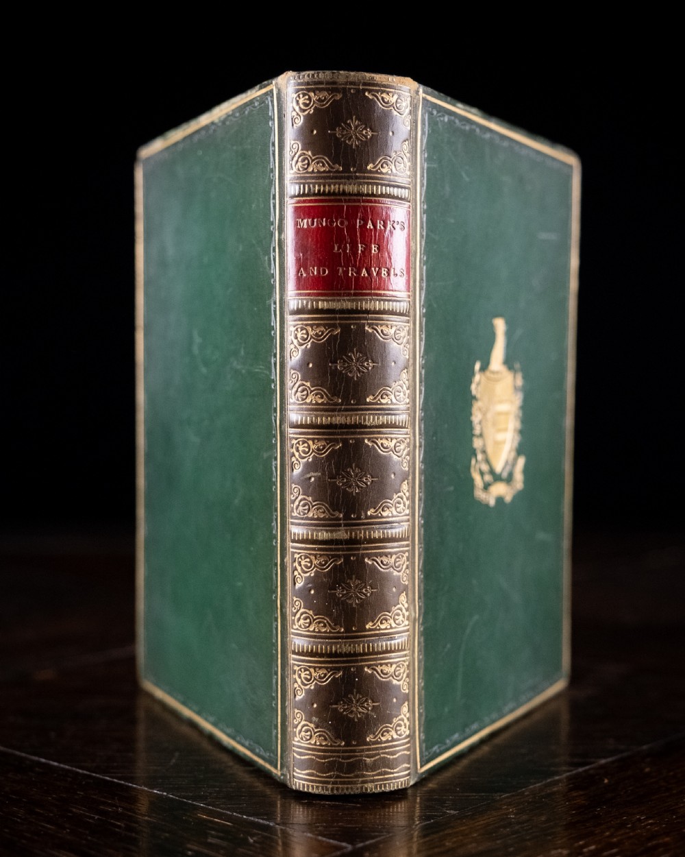 A Victorian leather-bound book standing upright, featuring green boards with a gilt crest and a brown leather spine decorated with gold tooling.
