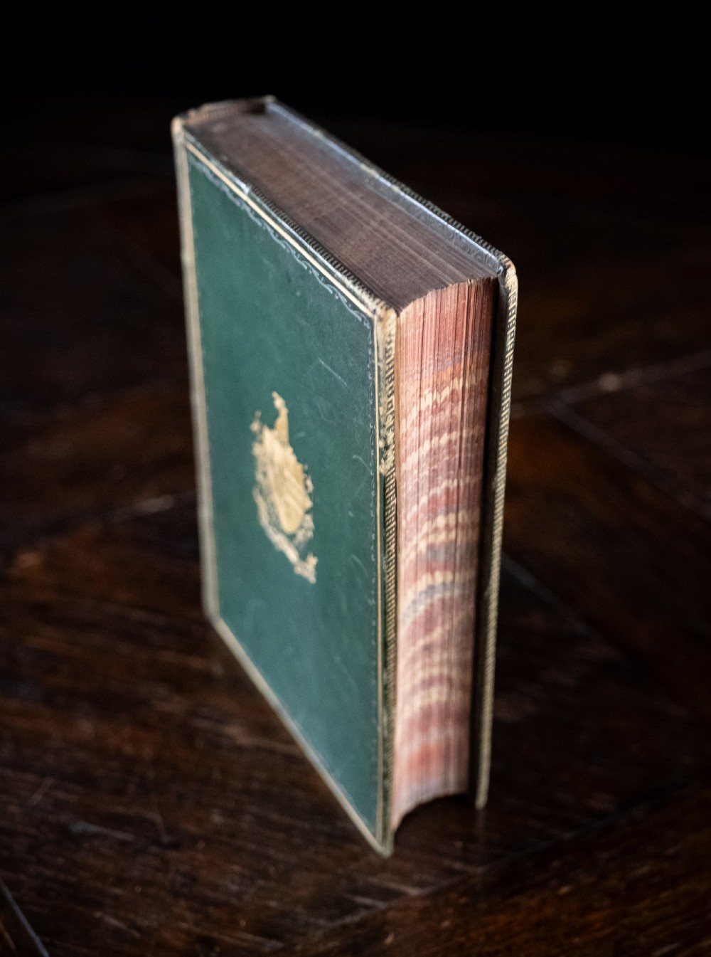 A Victorian leather-bound book standing upright, featuring green boards with a gilt crest and a brown leather spine decorated with gold tooling. View V