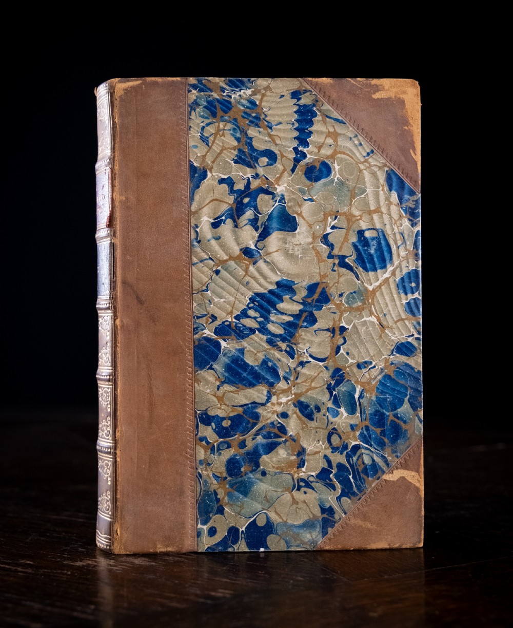 An 1864 half leather bound book featuring a brown gold-tooled spine with raised bands and blue marbled paper boards. View III