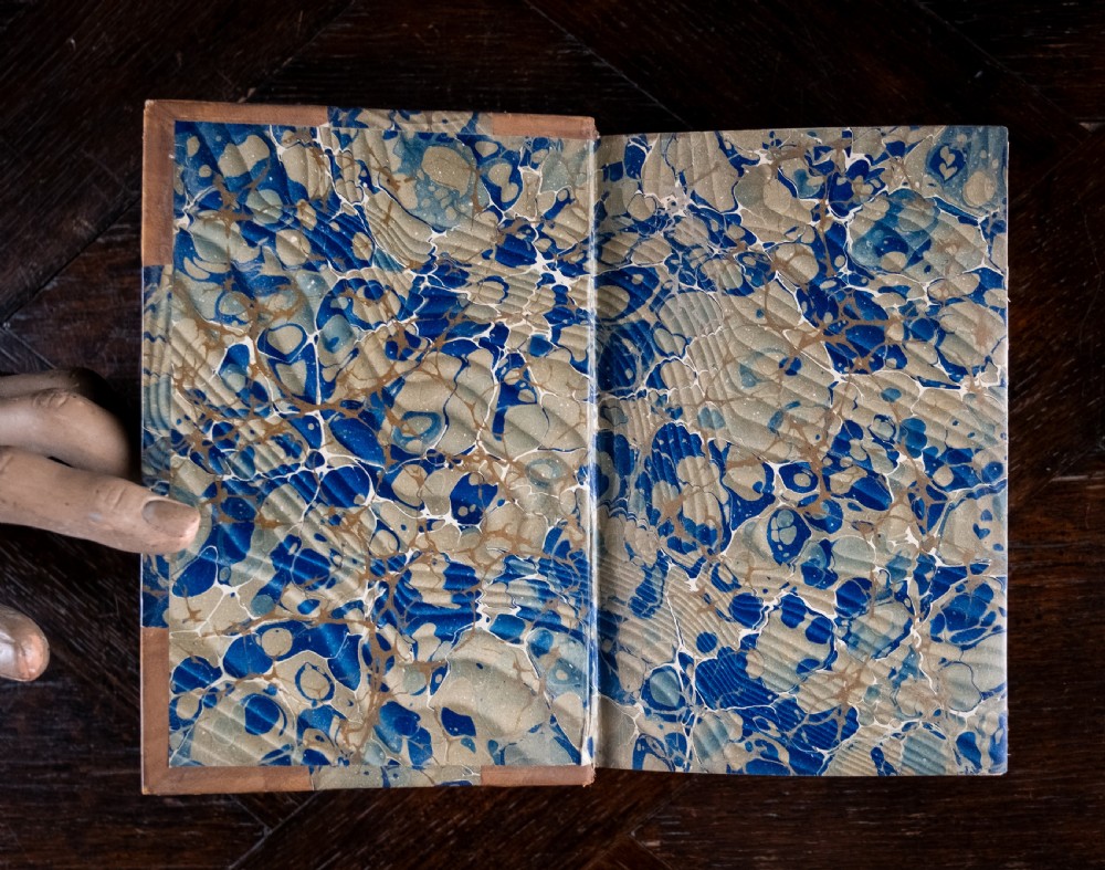 An 1864 half leather bound book featuring a brown gold-tooled spine with raised bands and blue marbled paper boards. View VII
