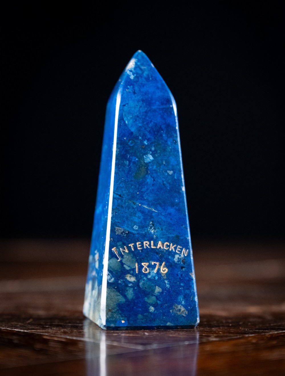 A polished deep blue lapis lazuli obelisk engraved with gilt text reading Interlacken 1876 near the base.
