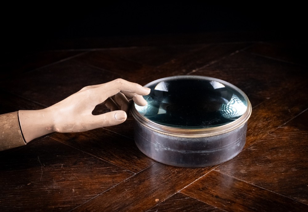 A Victorian cylindrical desktop magnifier featuring a thick circular glass convex lens set within a heavy metal rim.