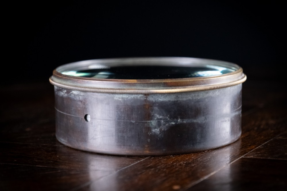 A Victorian cylindrical desktop magnifier featuring a thick circular glass convex lens set within a heavy metal rim. View V