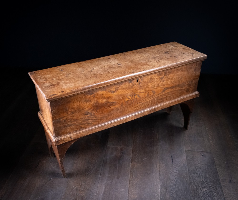 A seventeenth-century elm six-plank coffer featuring a hinged lid, raised bootjack side ends, and visible grain patterns against a dark background. View V