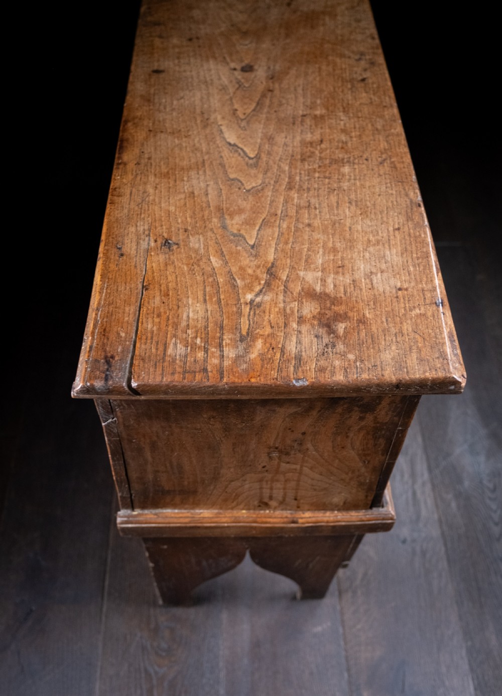 A seventeenth-century elm six-plank coffer featuring a hinged lid, raised bootjack side ends, and visible grain patterns against a dark background. View VI