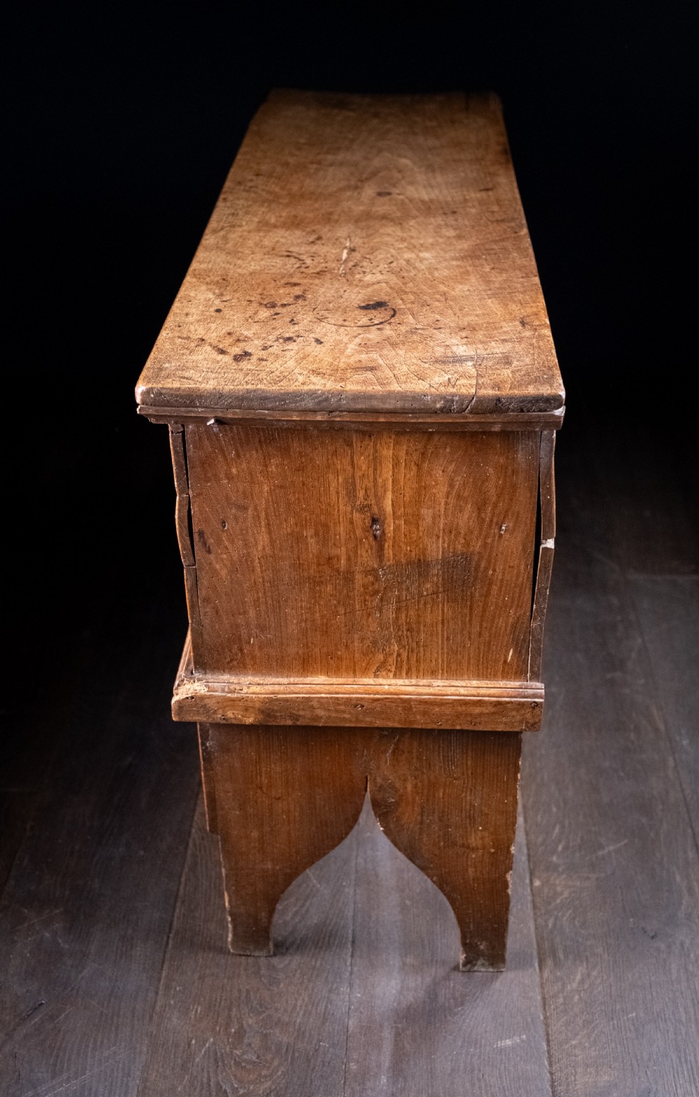 A seventeenth-century elm six-plank coffer featuring a hinged lid, raised bootjack side ends, and visible grain patterns against a dark background. View XVI