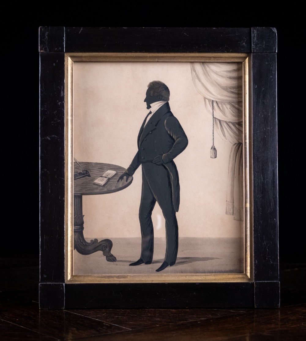 A framed full-length Regency silhouette portrait depicting a gentleman in profile standing beside a table with a draped curtain in the background.