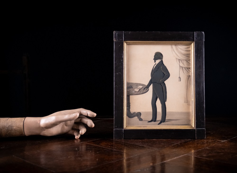 A framed full-length Regency silhouette portrait depicting a gentleman in profile standing beside a table with a draped curtain in the background. View II