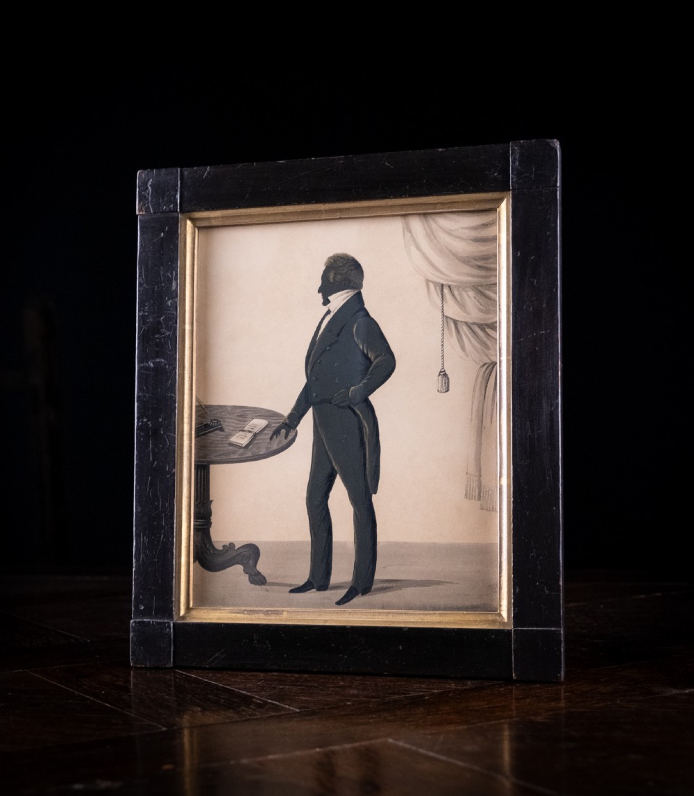 A framed full-length Regency silhouette portrait depicting a gentleman in profile standing beside a table with a draped curtain in the background. View V