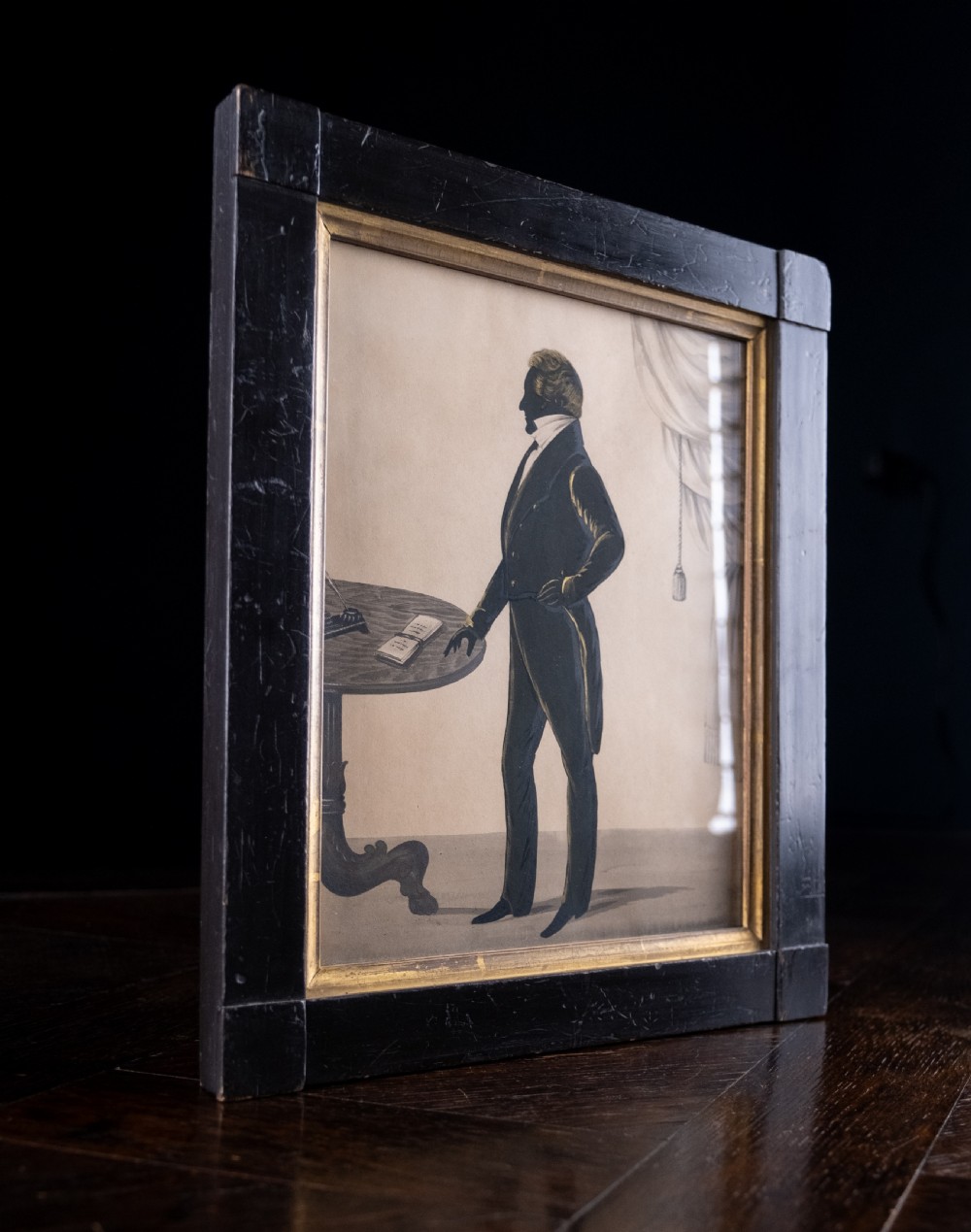 A framed full-length Regency silhouette portrait depicting a gentleman in profile standing beside a table with a draped curtain in the background. View VI