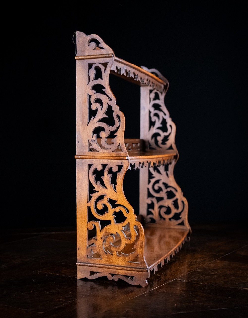 A three-tier Victorian mahogany wall shelf featuring open fretwork sides, pierced scroll decoration, and serrated apron trim. View V