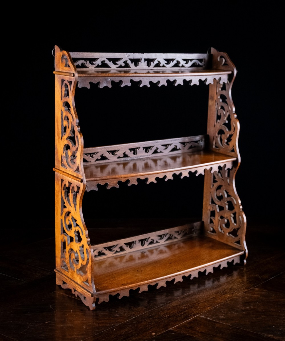 A three-tier Victorian mahogany wall shelf featuring open fretwork sides, pierced scroll decoration, and serrated apron trim. View VI