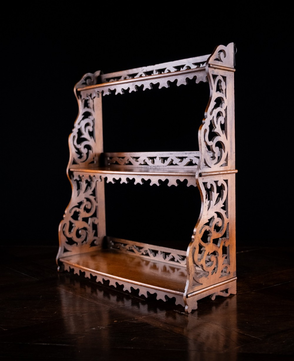 A three-tier Victorian mahogany wall shelf featuring open fretwork sides, pierced scroll decoration, and serrated apron trim. View VII