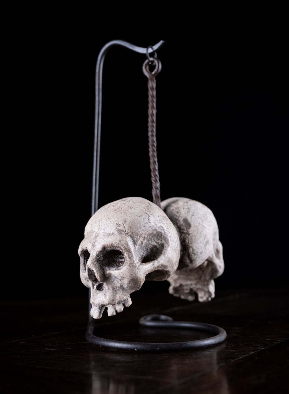A pair of small carved off-white skull sculptures suspended from a twisted metal chain attached to a black wrought iron stand.