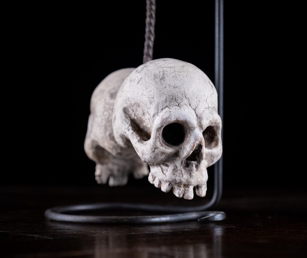 A pair of small carved off-white skull sculptures suspended from a twisted metal chain attached to a black wrought iron stand. View III