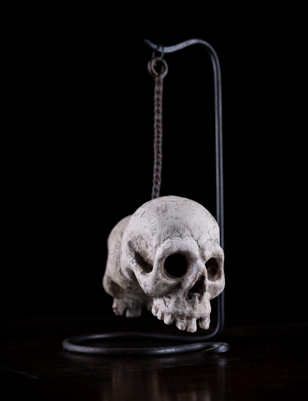 A pair of small carved off-white skull sculptures suspended from a twisted metal chain attached to a black wrought iron stand. View IV