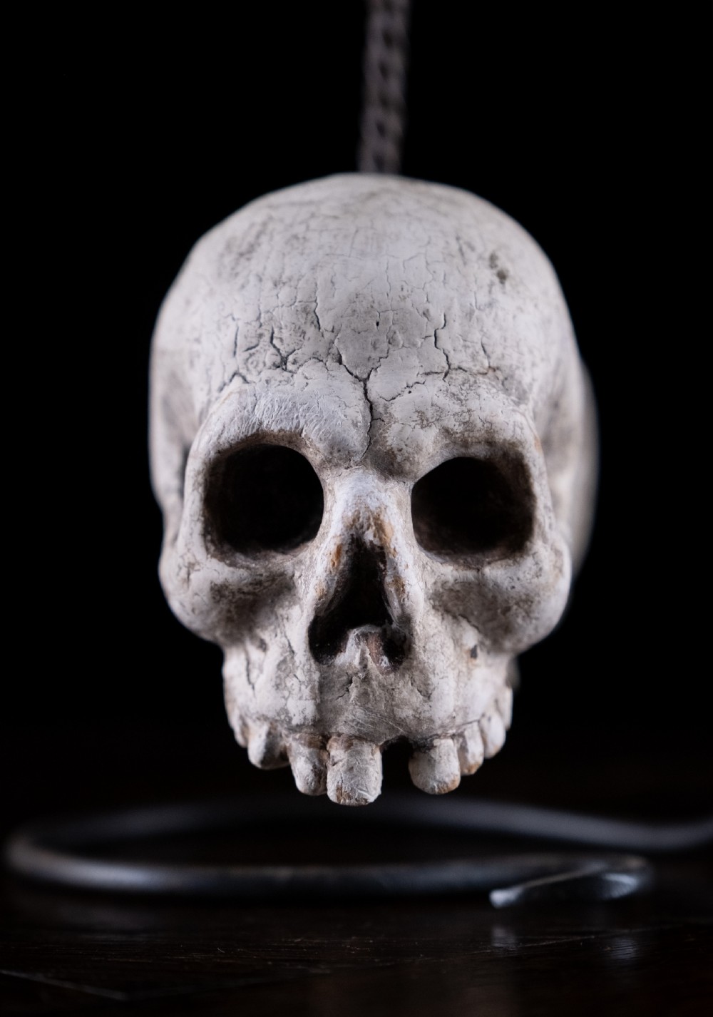 A pair of small carved off-white skull sculptures suspended from a twisted metal chain attached to a black wrought iron stand. View VI