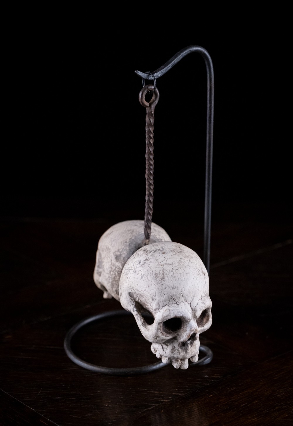 A pair of small carved off-white skull sculptures suspended from a twisted metal chain attached to a black wrought iron stand. View VII