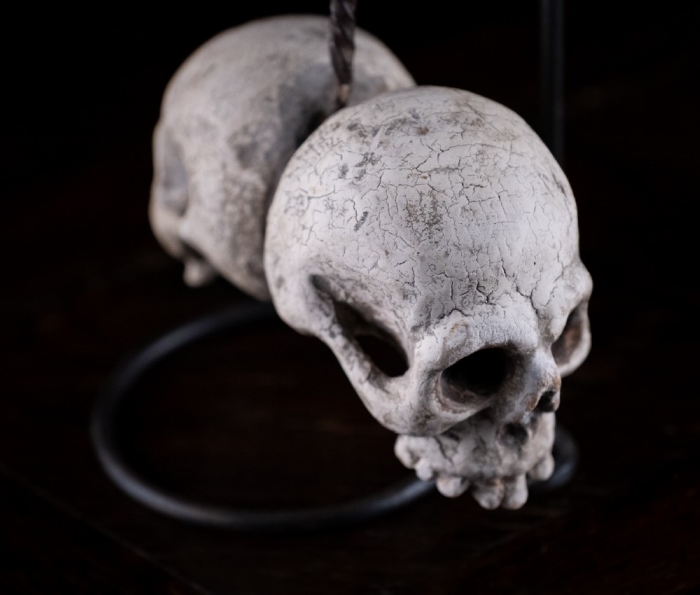 A pair of small carved off-white skull sculptures suspended from a twisted metal chain attached to a black wrought iron stand. View VIII