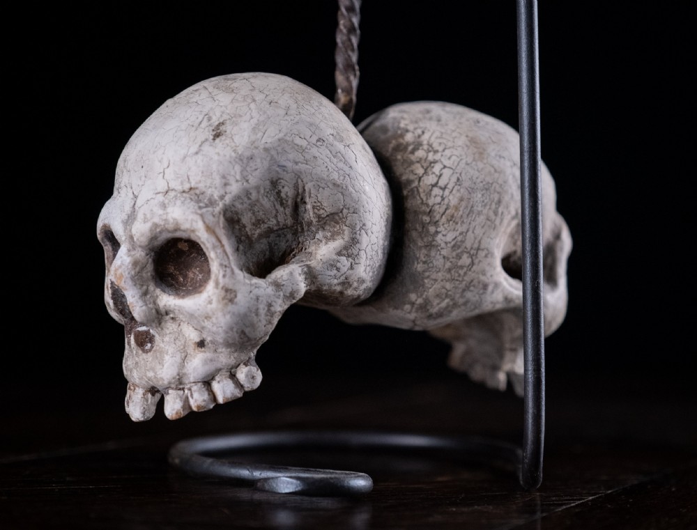A pair of small carved off-white skull sculptures suspended from a twisted metal chain attached to a black wrought iron stand. View IX