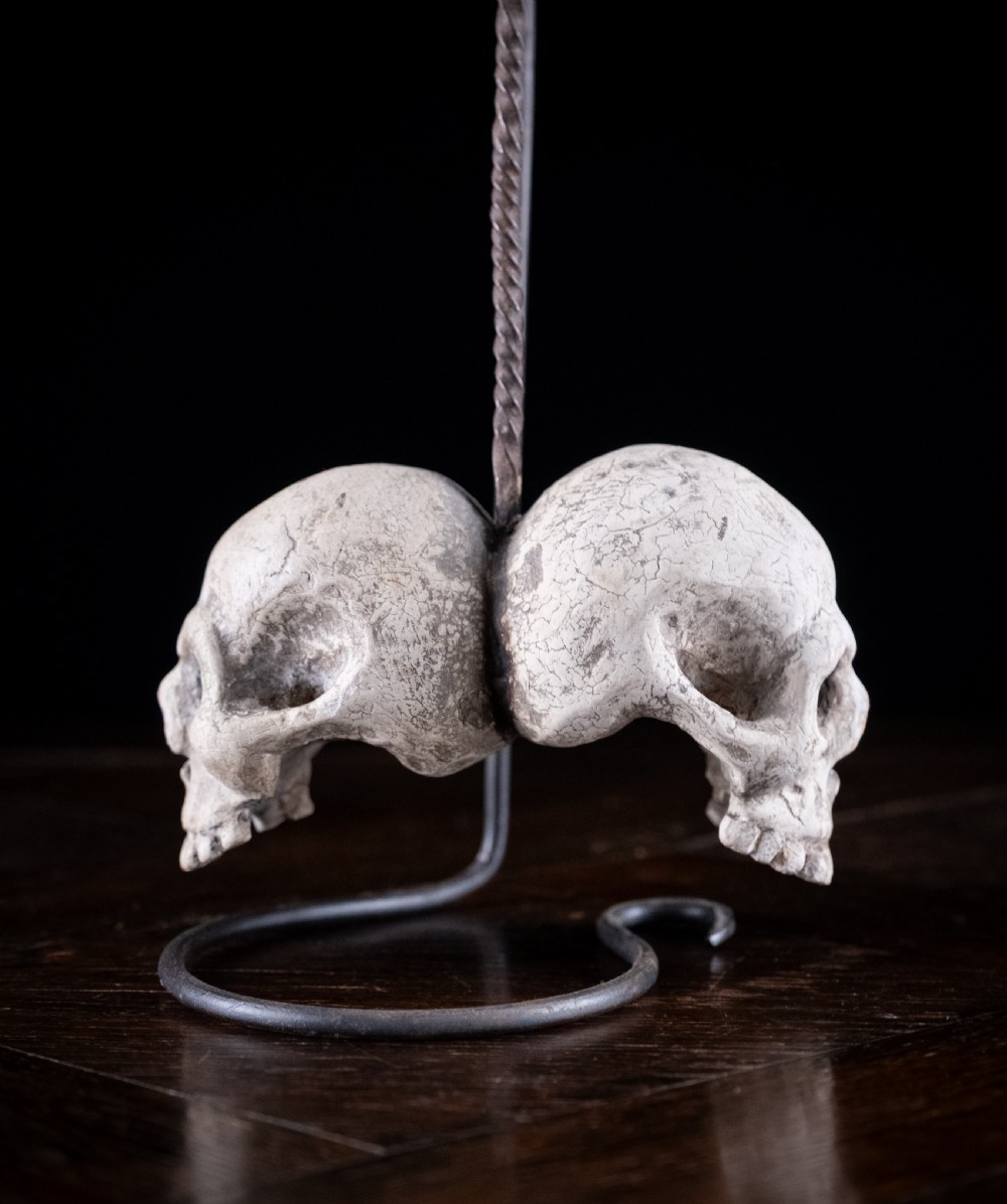 A pair of small carved off-white skull sculptures suspended from a twisted metal chain attached to a black wrought iron stand. View X