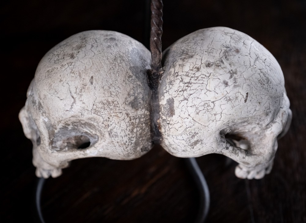 A pair of small carved off-white skull sculptures suspended from a twisted metal chain attached to a black wrought iron stand. View XI