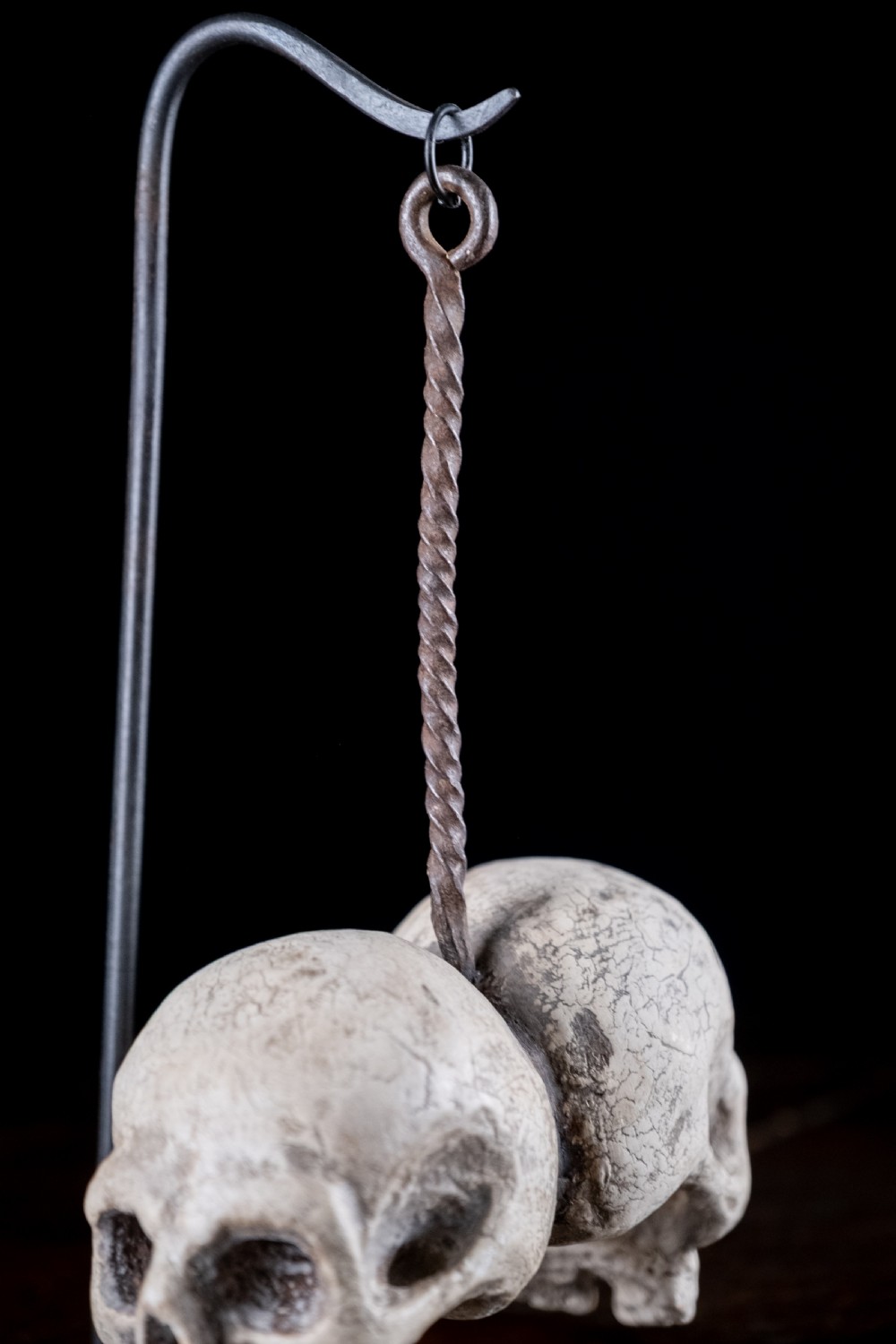 A pair of small carved off-white skull sculptures suspended from a twisted metal chain attached to a black wrought iron stand. View XII