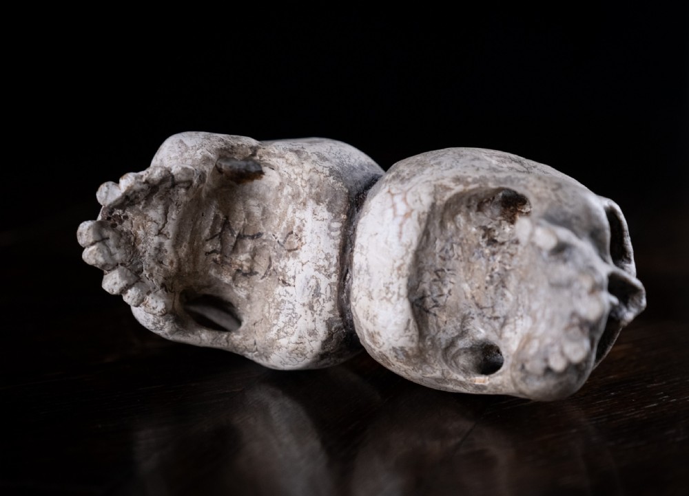 A pair of small carved off-white skull sculptures suspended from a twisted metal chain attached to a black wrought iron stand. View XIII