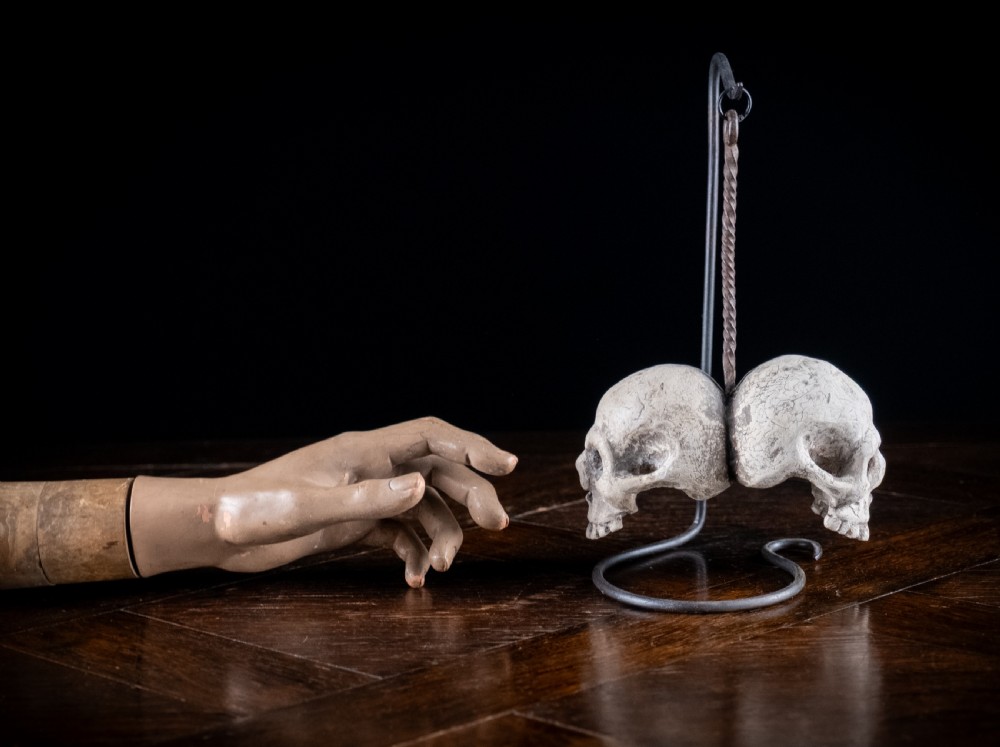 A pair of small carved off-white skull sculptures suspended from a twisted metal chain attached to a black wrought iron stand. View XVI