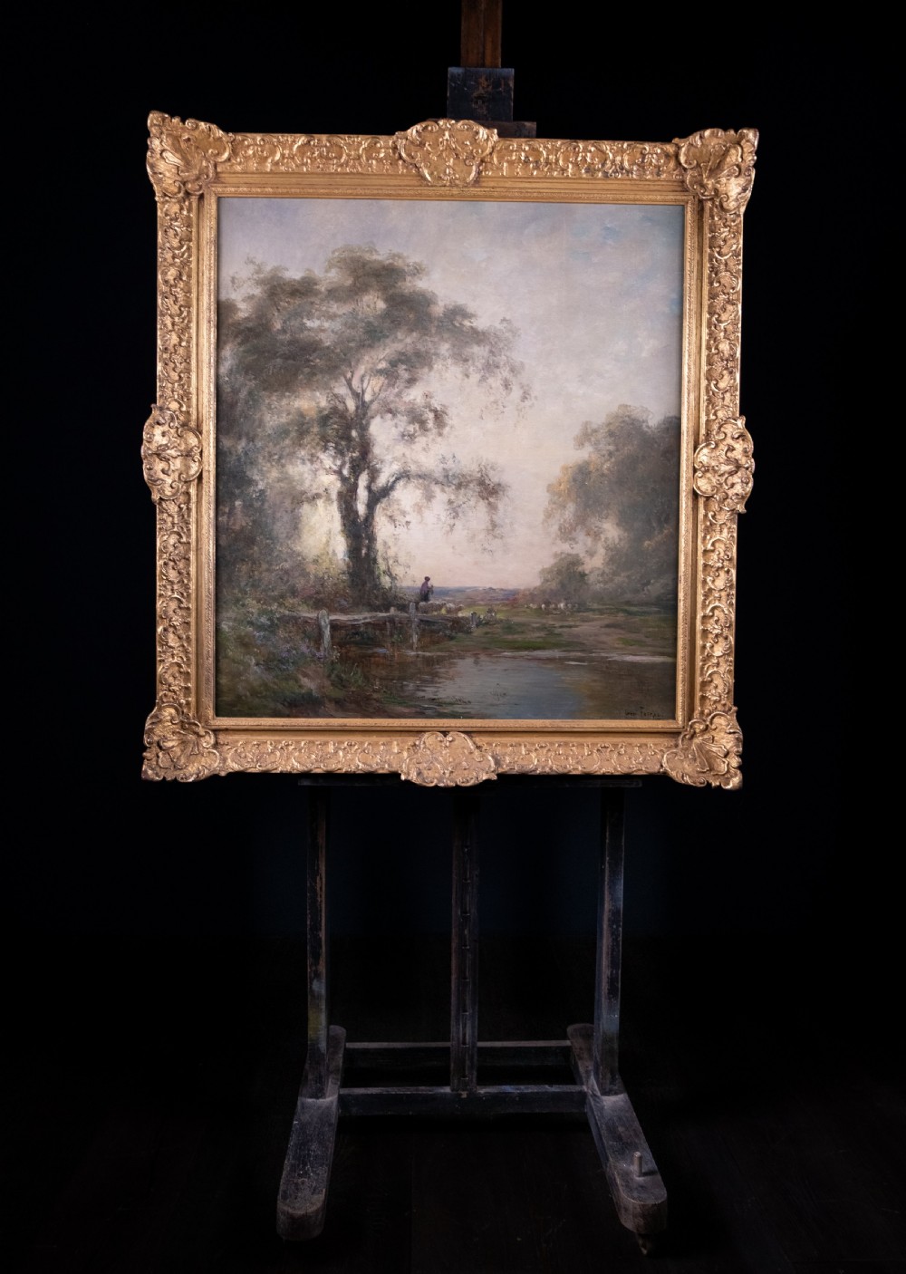 An impressionist oil painting depicting a rural countryside landscape with a large tree by a river, enclosed within an ornate gilded frame. View II