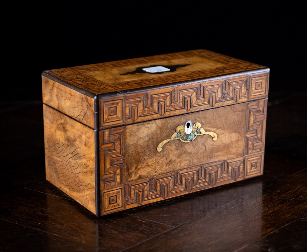 A rectangular walnut tea caddy featuring geometric parquetry borders, a mother-of-pearl escutcheon, and brass inlay on a dark wooden surface.