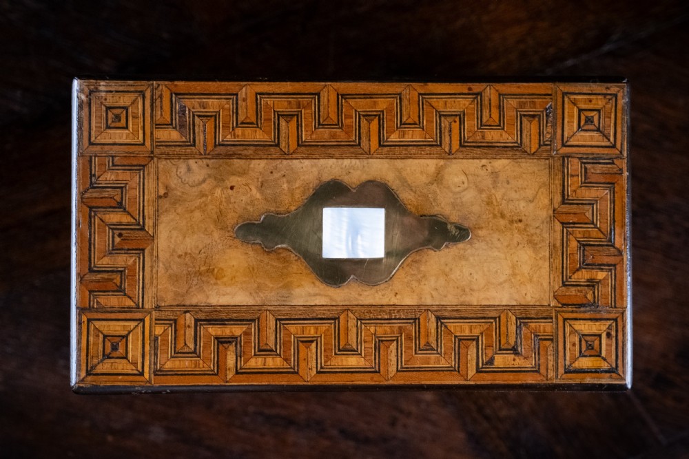 A rectangular walnut tea caddy featuring geometric parquetry borders, a mother-of-pearl escutcheon, and brass inlay on a dark wooden surface. View V