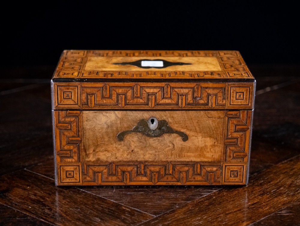 A rectangular walnut tea caddy featuring geometric parquetry borders, a mother-of-pearl escutcheon, and brass inlay on a dark wooden surface. View VI