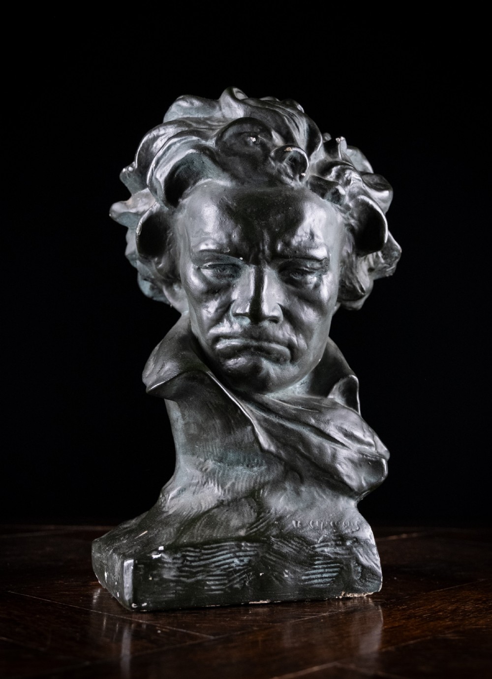 A dark bronze-coloured painted plaster bust depicting composer Ludwig van Beethoven with detailed hair and a dramatic collar on a square base. View II