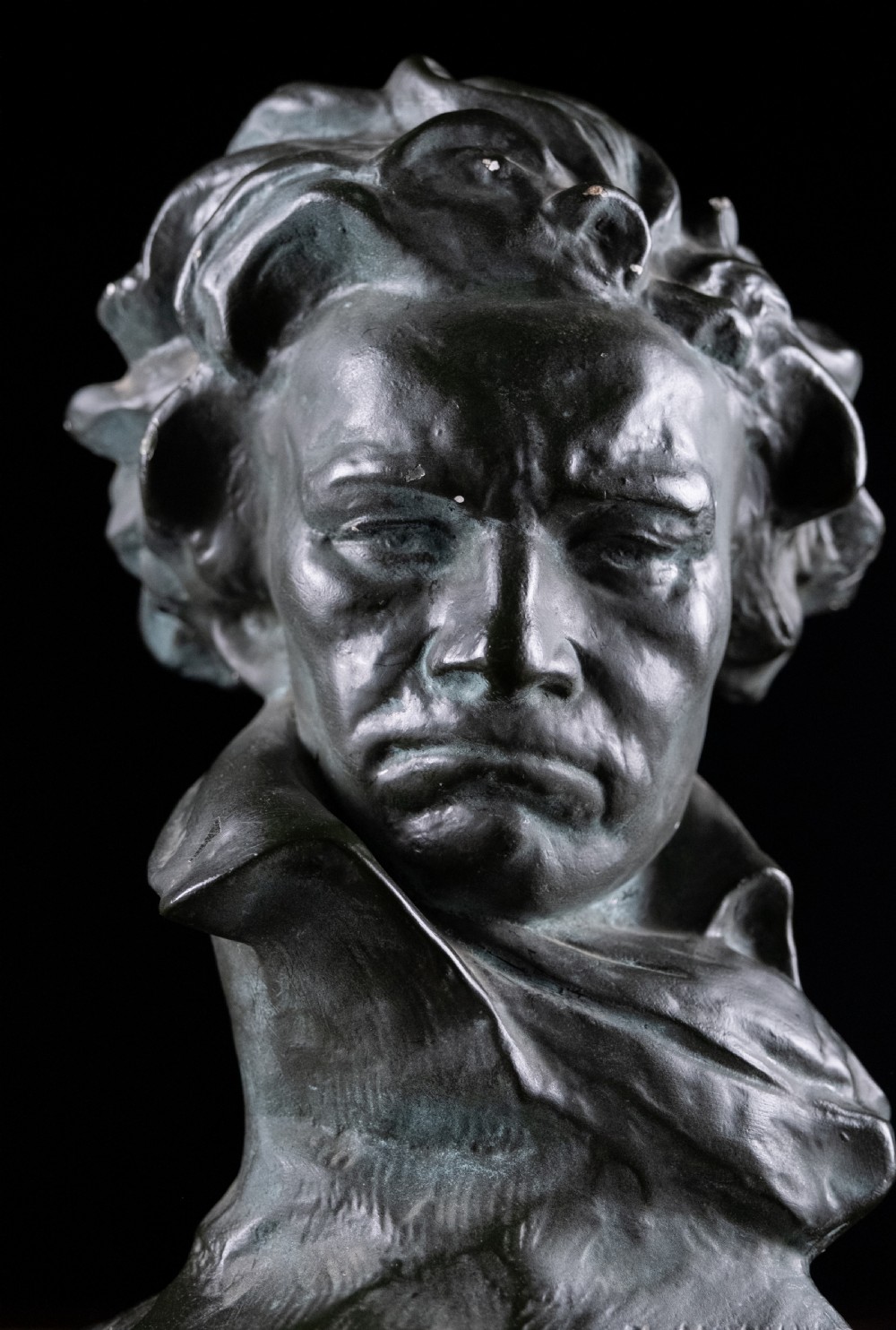 A dark bronze-coloured painted plaster bust depicting composer Ludwig van Beethoven with detailed hair and a dramatic collar on a square base. View III