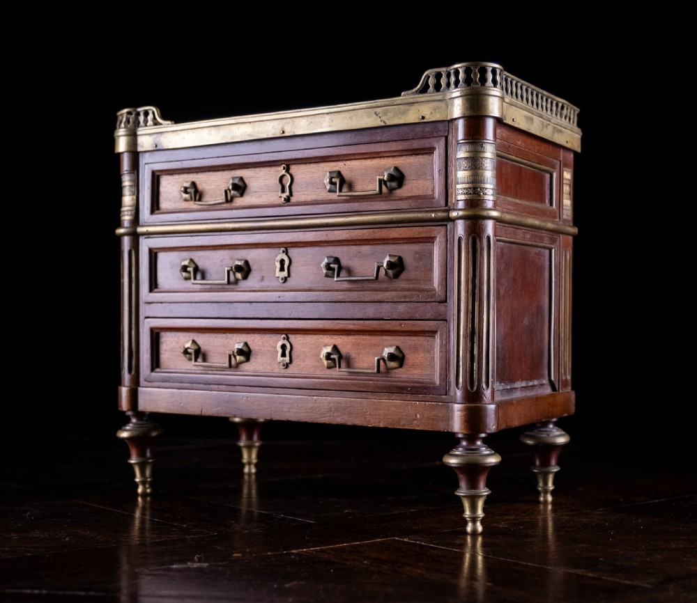 A miniature Louis XVI style mahogany three-drawer commode featuring brass mounts, fluted corner columns, and a gallery top inset with marble. View IV