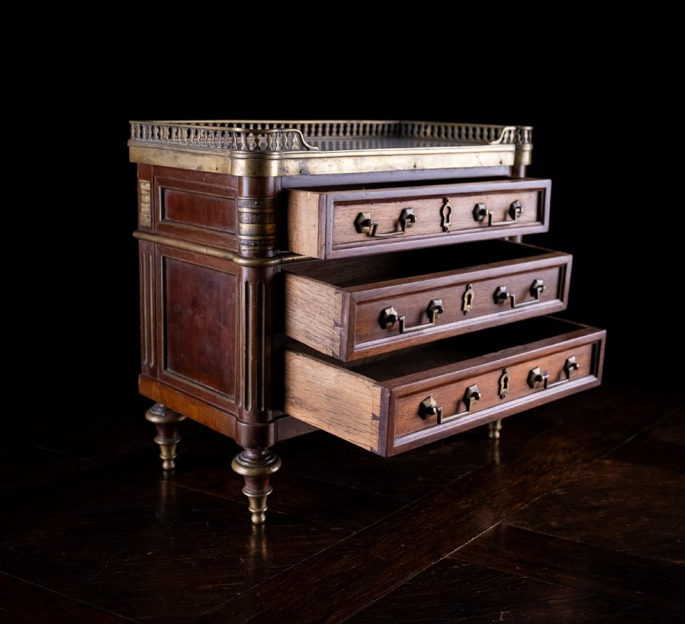 A miniature Louis XVI style mahogany three-drawer commode featuring brass mounts, fluted corner columns, and a gallery top inset with marble. View VI