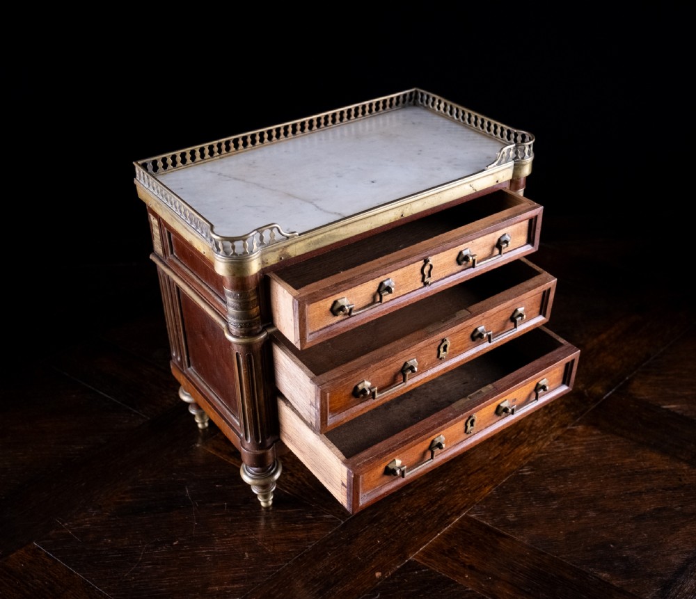 A miniature Louis XVI style mahogany three-drawer commode featuring brass mounts, fluted corner columns, and a gallery top inset with marble. View VIII