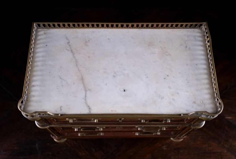 A miniature Louis XVI style mahogany three-drawer commode featuring brass mounts, fluted corner columns, and a gallery top inset with marble. View IX
