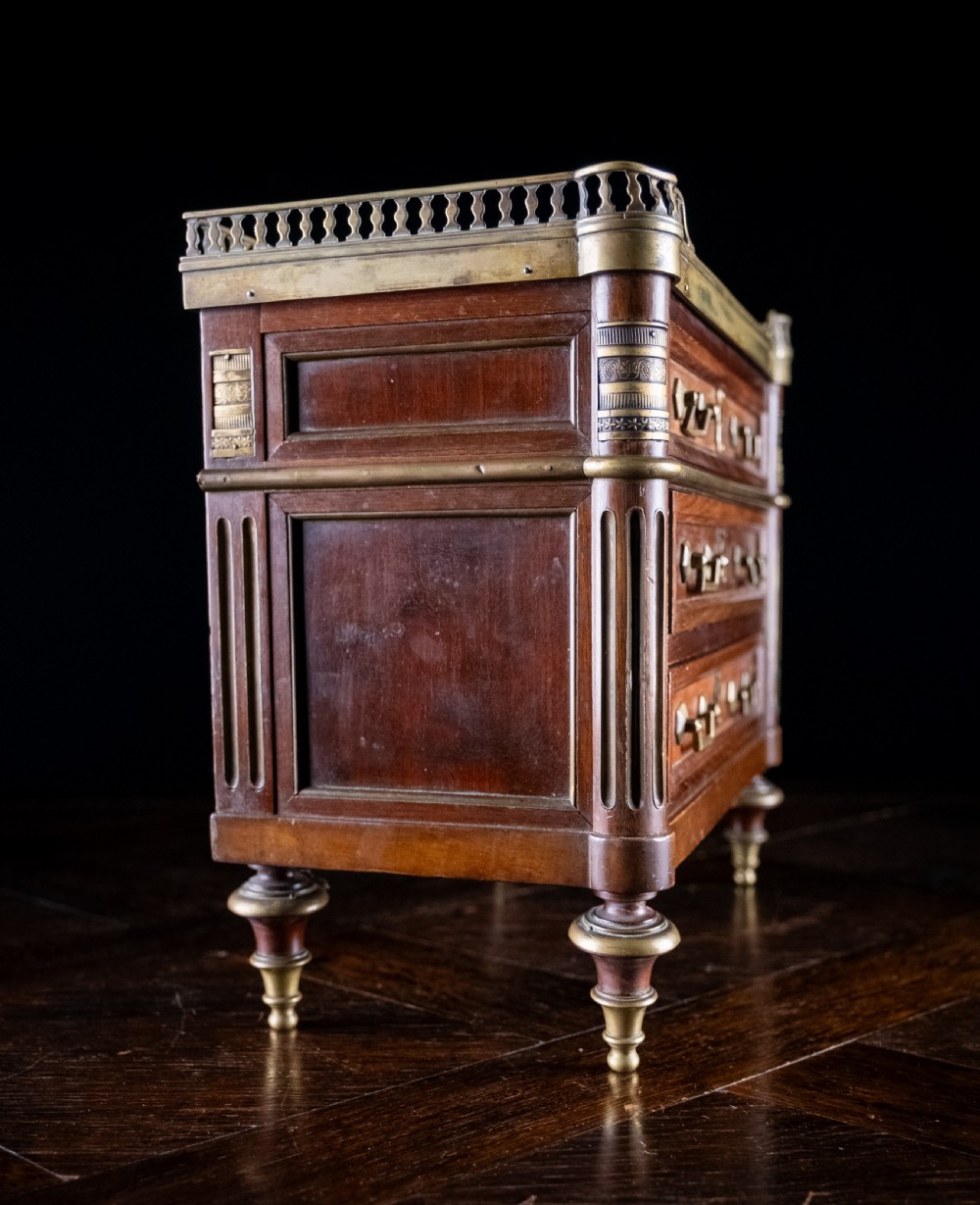 A miniature Louis XVI style mahogany three-drawer commode featuring brass mounts, fluted corner columns, and a gallery top inset with marble. View XX