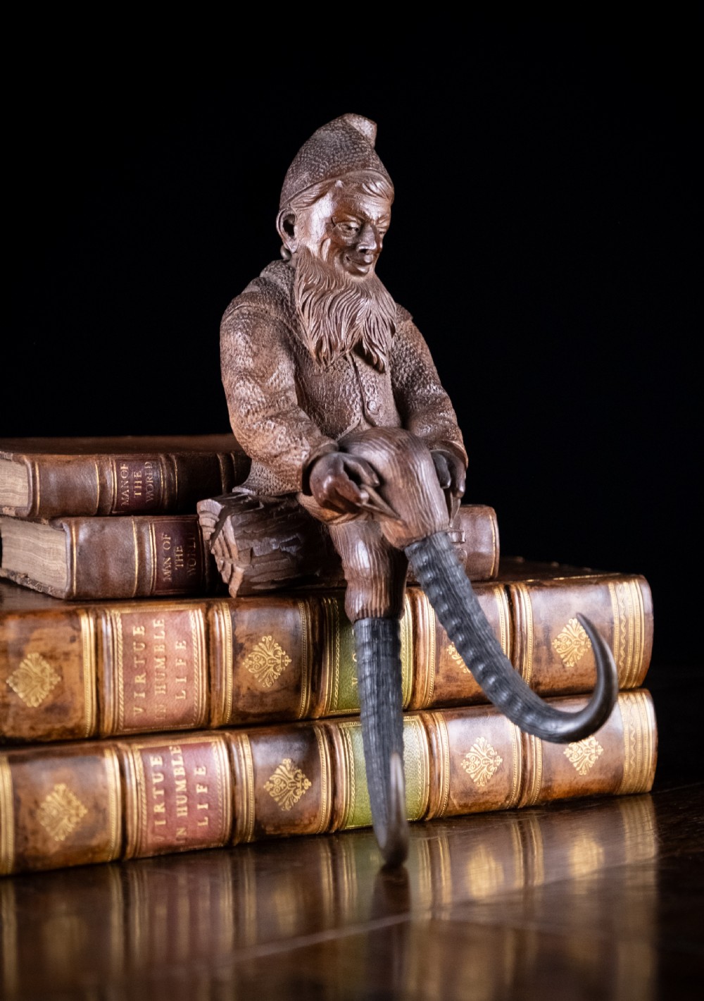 A dark carved wooden Black Forest gnome figure seated on a log with curved horn hooks extending as legs.