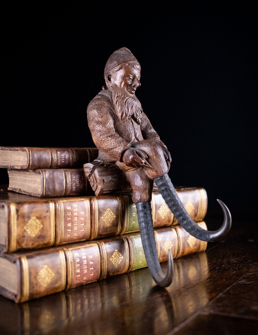 A dark carved wooden Black Forest gnome figure seated on a log with curved horn hooks extending as legs. View II