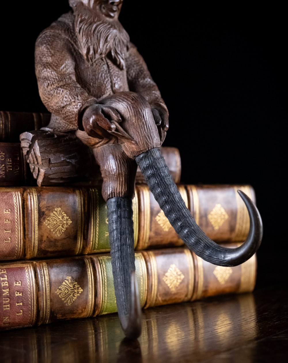A dark carved wooden Black Forest gnome figure seated on a log with curved horn hooks extending as legs. View III