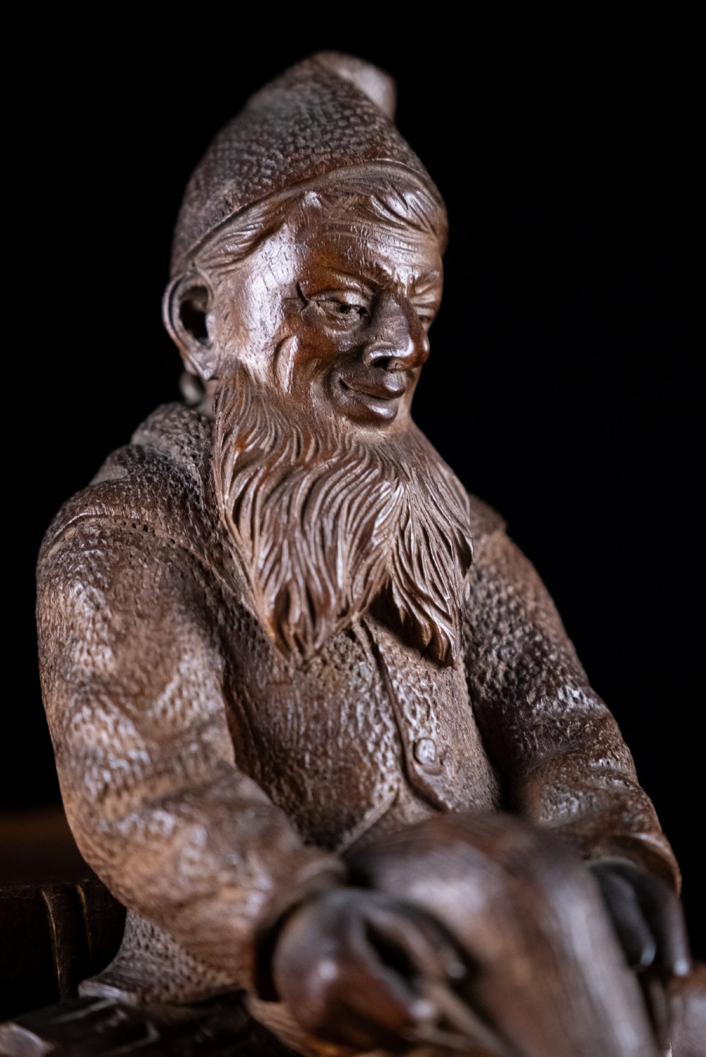 A dark carved wooden Black Forest gnome figure seated on a log with curved horn hooks extending as legs. View IV