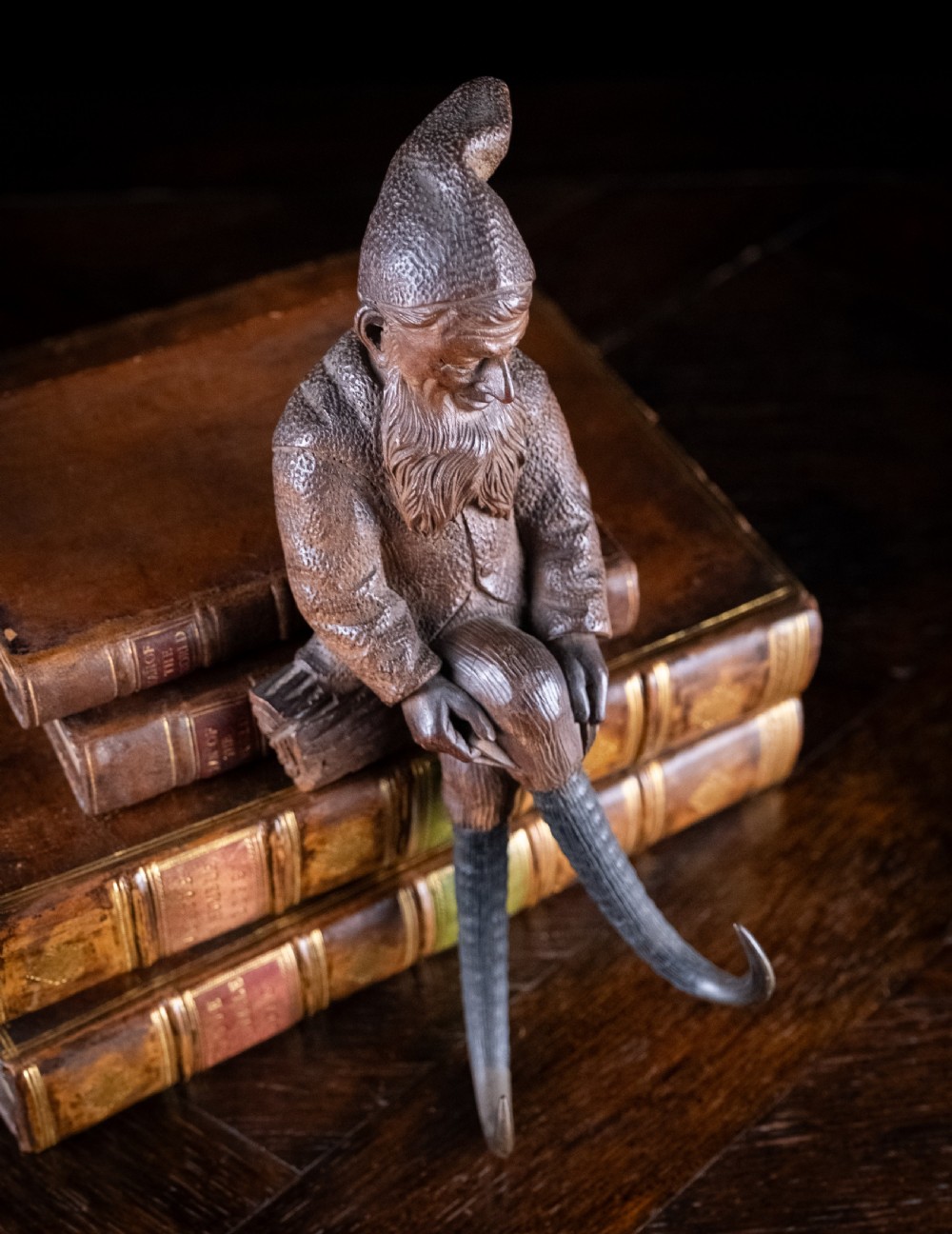 A dark carved wooden Black Forest gnome figure seated on a log with curved horn hooks extending as legs. View X