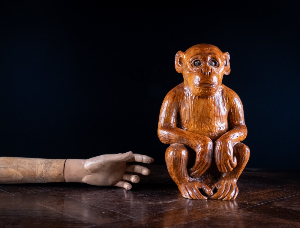 A hand-carved warm brown wooden sculpture depicting a seated monkey with chiselled fur details and dark eyes. View II