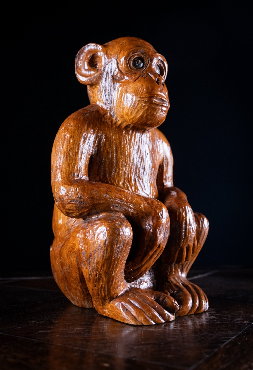 A hand-carved warm brown wooden sculpture depicting a seated monkey with chiselled fur details and dark eyes. View III