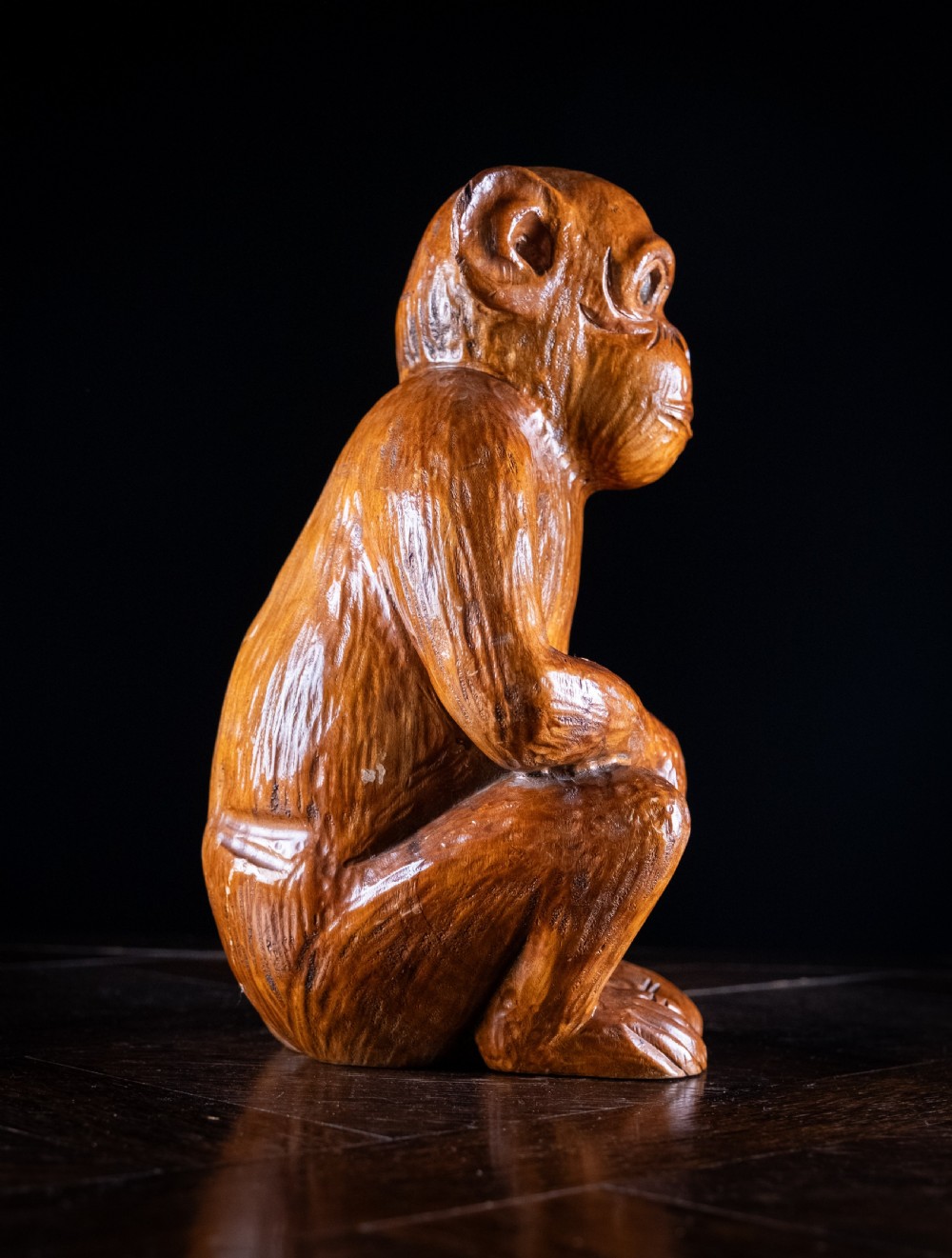 A hand-carved warm brown wooden sculpture depicting a seated monkey with chiselled fur details and dark eyes. View IV