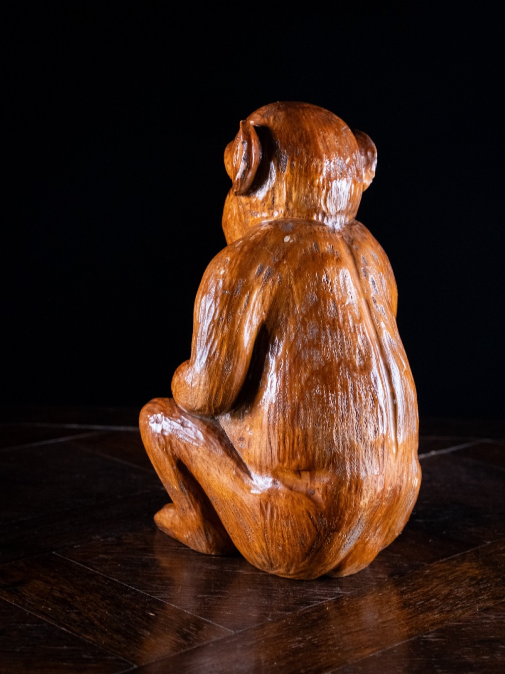 A hand-carved warm brown wooden sculpture depicting a seated monkey with chiselled fur details and dark eyes. View VII