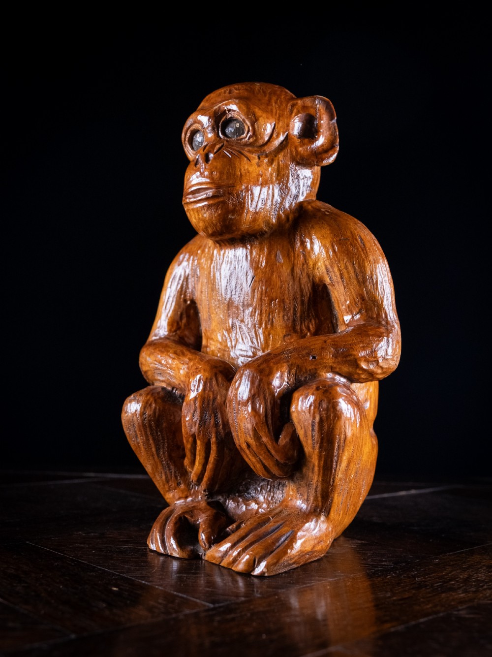 A hand-carved warm brown wooden sculpture depicting a seated monkey with chiselled fur details and dark eyes. View VIII
